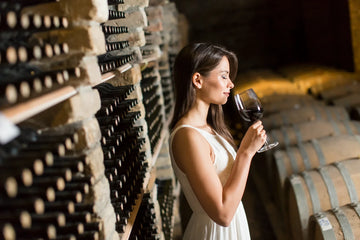 Woman holding a glass of red wine in a wine cellar with rows of wine bottles and barrels.