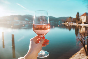 Hand holding a glass of rose wine by a lake with mountains in the background