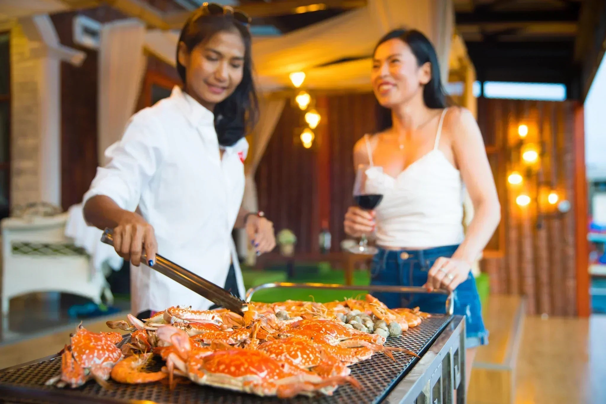 Two Asian women enjoying wine together while grilling fresh prawns on an outdoor table, sharing food and conversation in a relaxed and warm atmosphere.